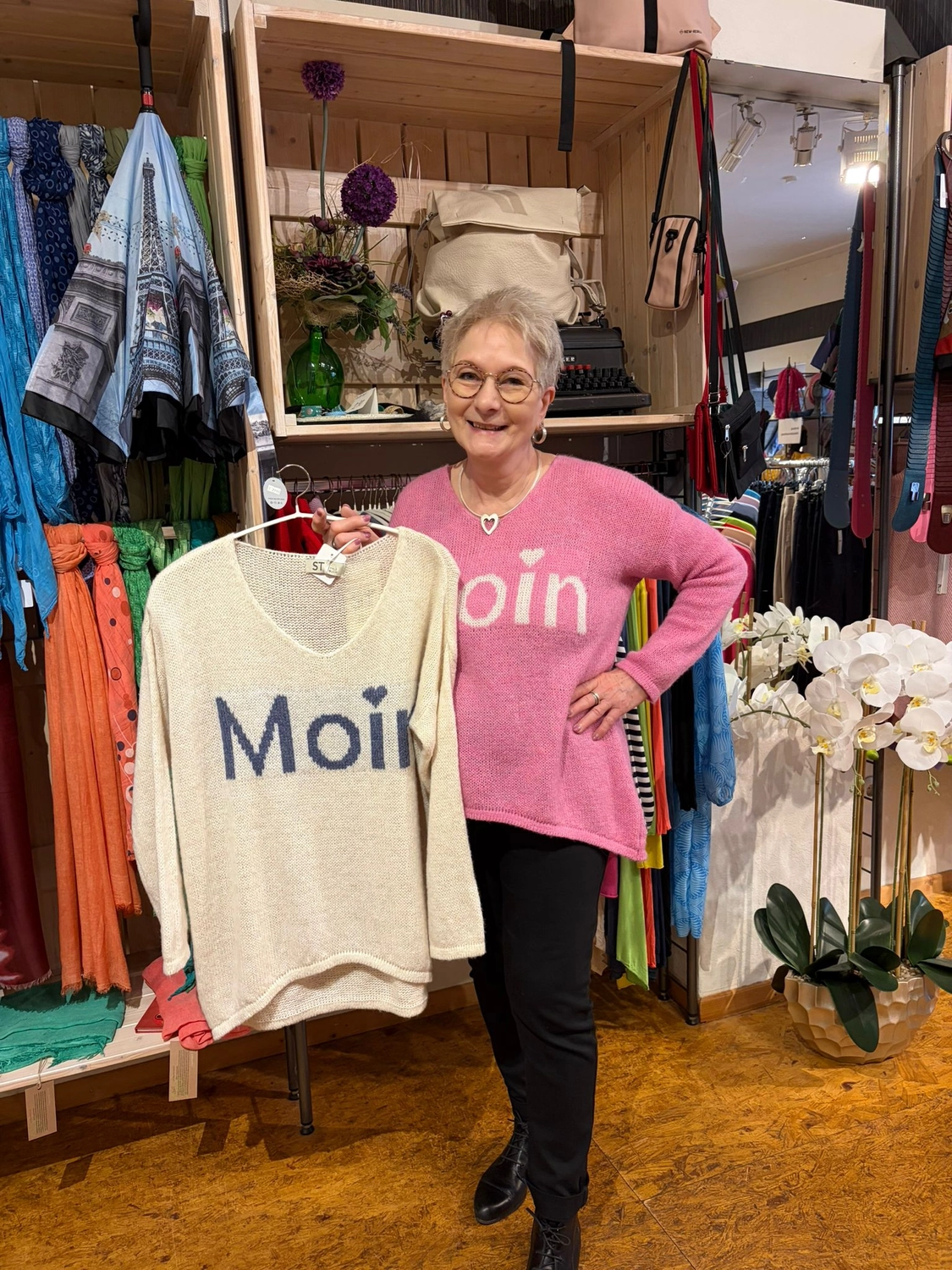 Mode & Handarbeitsdiele - Wolle, Kunsthandwerk, Mode und Accessoires - Eine lächelnde Frau mit Brille und einem rosa „Moon“-Pullover hält in einer Boutique ein cremefarbenes Stricktop hoch.